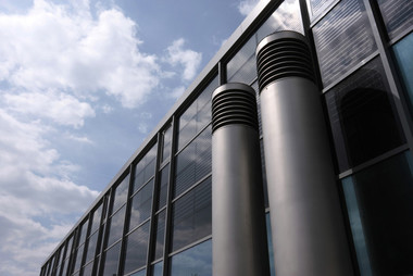 Two tall cylindrical metal vents on the exterior of a glass-clad building, with a blue sky and clouds in the background.