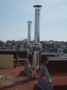 Two tall white chimneys rise from a red rooftop with a city skyline in the distance.