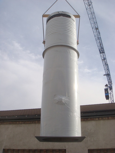 A large white cylindrical storage silo is suspended by a crane above a building’s roof.