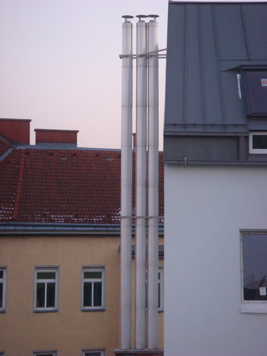 Three tall silver chimneys rise above the rooftops between a yellow building on the left and a white building on the right.