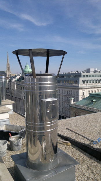 Shiny stainless-steel rooftop exhaust vent with a circular cap on a flat roof, city skyline in the distance.