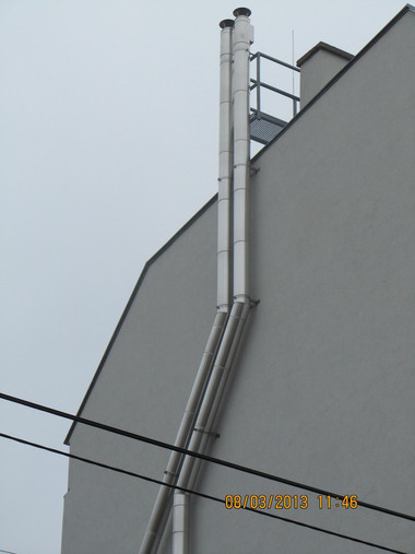 Side view of a gray building wall with a vertical metal vent stack and diagonal ducts, with power lines crossing the foreground.