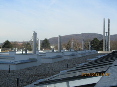 Flat rooftop with numerous vertical vent pipes and skylights, gravel surface, and a distant hill under a blue sky.
