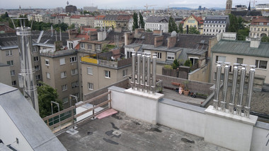 Rooftop terrace overlooking a dense urban neighborhood of low-rise buildings with chimneys and a distant city skyline.