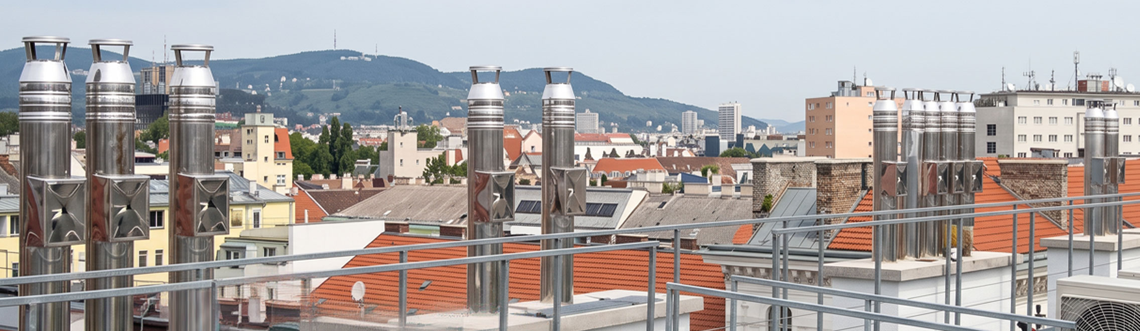 moldrich-26-produkte-header-01 Rooftop view with numerous metal vent stacks overlooking a city of red-tiled roofs and distant hills.