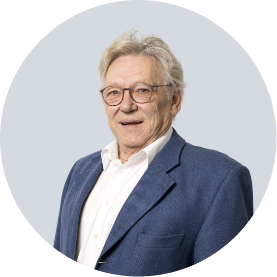 Portrait of an elderly man wearing glasses, a white shirt, and a blue blazer, smiling against a light blue background.
